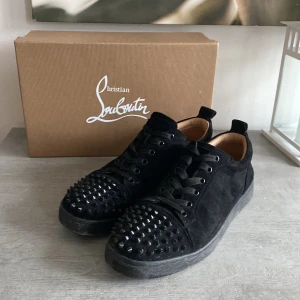 Svarta Louboutins - Snygga svarta sneakers i mocka från Christian Louboutin med coola nitar på tån och klassisk röd sula. Skorna har snörning och en stilren design som sticker ut.