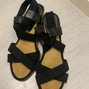 Svarta sandaler med kilklack - Snygga svarta sandaler med kilklack och flätad sula. De har breda remmar i textil och en elastisk hälrem för extra komfort. Perfekta för sommarens varma dagar. Från din sko. AI valt annat märke som inte kan tas bort.
