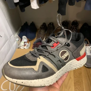 Grå sneakers från Colmar - Snygga grå sneakers från Colmar med beige och svarta detaljer. Skorna har en robust sula med röda inslag och snörning framtill. Perfekta för en stilren och bekväm look.