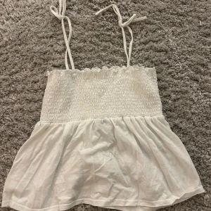 Vit ärmlös topp från Vero Moda - Säljer en söt vit ärmlös topp från Vero Moda med smockdetaljer och tunna axelband som knyts. Perfekt för varma sommardagar. Toppen har en luftig och lätt design. Storlek M