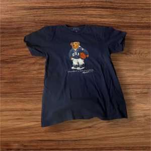 Mörkblå t-shirt med björntryck från Polo Ralph Lauren - Säljer en mörkblå polo bear tshirt. . T-shirten är kortärmad och tillverkad i 100% bomull, vilket gör den bekväm och lätt att bära. Storleken passade inte och därför säljer jag.