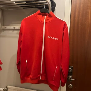 Röd track jacket från Palm Angels - Snygg röd track jacket från Palm Angels med vita detaljer och dragkedja. Jackan har långa ärmar med vita och svarta ränder längs sidorna och två fickor med dragkedja framtill. Säljer av hela garderoben då kläderna inte används längre.