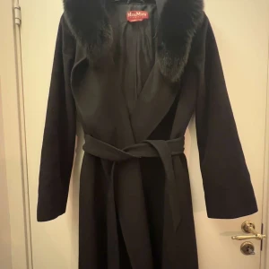 Svart kappa från MaxMara - Elegant svart kappa från MaxMara med en lyxig pälskrage och knytbälte i midjan. Tillverkad i ren ny ull för en varm och stilren look. Perfekt för kyliga dagar när du vill känna dig både bekväm och chic.