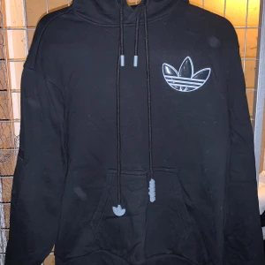Svart hoodie från Adidas - Säljer en svart hoodie från Adidas med det klassiska trefoil-logotypen på bröstet. Hoodien har en justerbar dragsko i huvan och en stor magficka framtill. Perfekt för en avslappnad stil. 