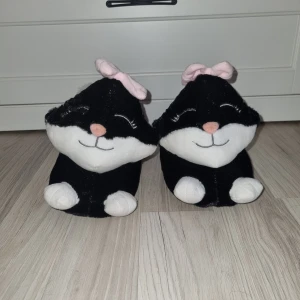 Svarta tofflor med kattdesign - Mysiga svarta tofflor formade som katter med vita detaljer och rosa rosetter. De har en söt design med små öron och svansar, perfekt för att hålla fötterna varma hemma. Sulan är svarta med halkskydd för extra säkerhet.