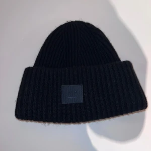 Svart mössa från Acne Studios - Endast testad!! Snygg svart ribbad mössa från Acne Studios med ett stilrent ansiktsmärke framtill. Perfekt för att hålla värmen under kyliga dagar. Mössan är tillverkad i ett mjukt och bekvämt material. 