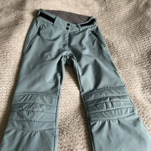 Gröna täckbyxor från McKinley - Snygga gröna täckbyxor från McKinley med dragkedjor vid fickorna och knappar i midjan. Perfekta för vinterns äventyr med förstärkta knän för extra hållbarhet. Byxorna har en bekväm passform och är idealiska för kalla dagar.