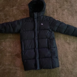 Svart dunjacka från Calvin Klein - Snygg svart dunjacka från Calvin Klein med lång ärm och hög krage. Jackan har en stilren design med diskret CK-logga på bröstet och är perfekt för kyliga dagar. Den är quiltad för extra värme och komfort.