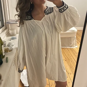 Vit klänning med broderade detaljer - Säljer en söt vit klänning med svarta broderade detaljer vid hals och ärmslut. Klänningen har långa ärmar och en lös passform, perfekt för en avslappnad stil. Går att klä upp med ett coolt skärp! 