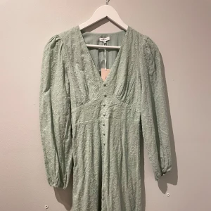 Ljusgrön långklänning med knappar - Charmig ljusgrön långklänning med v-ringning och knappar framtill. Klänningen har långa ärmar och ett diskret blommigt mönster. Perfekt för vår och sommar! Aldrig använd, lapp finns kvar.
