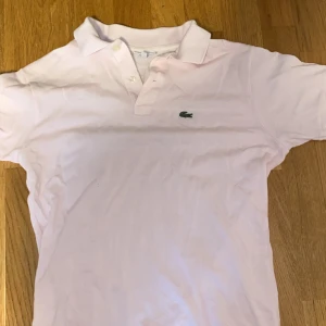 Ljusrosa pikétröja från Lacoste - Säljer en stilren ljusrosa pikétröja från Lacoste med klassisk krage och knappar framtill. Tröjan har det ikoniska krokodilmärket på bröstet. Perfekt för en avslappnad och snygg look.