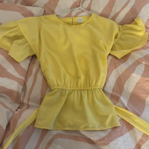 Gul blus från Lindex - Säljer en söt gul blus från Lindex med korta volangärmar och knytband i midjan. Perfekt för en färgglad look. Blusen har en elastisk midja för en bekväm passform. Blusen har en fläck på ärmen men går nog att tvätta bort!! ❤️