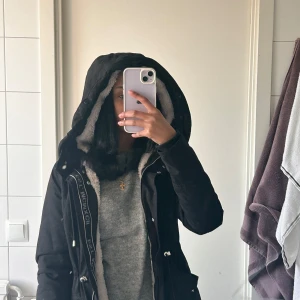 Svart parkajacka med foder - Säljer en svart parkajacka med mysigt foder i huvan. Jackan har dragkedja och knappar framtill samt snörning i midjan för en justerbar passform. Perfekt för kyliga dagar.