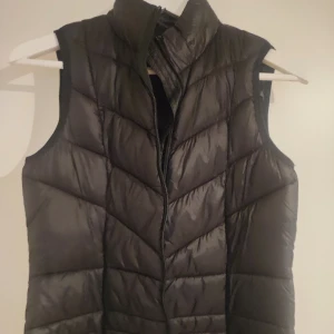 Svart dunväst från Vero Moda - Snygg svart dunväst från Vero Moda med dragkedja framtill. Perfekt för kyligare dagar och ger en stilren look. Västen har en quiltad design som ger extra värme och komfort.