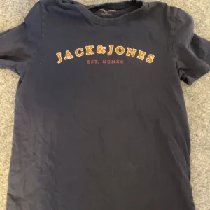 Mörkblå t-shirt från Jack & Jones - Säljer en mörkblå t-shirt från Jack & Jones med tryck på bröstet. Perfekt för en avslappnad stil. T-shirten är i bomull och har korta ärmar. Finns ett litet hål längst ner på tröjan!