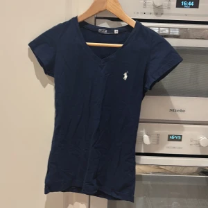 Mörkblå t-shirt från Polo - Säljer en mörkblå t-shirt från Ralph Lauren i 100% bomull. T-shirten har en v-ringning och korta ärmar med en liten broderad logotyp på bröstet. Perfekt för en avslappnad stil.