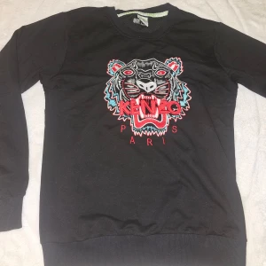 Svart sweatshirt från Kenzo - Säljer en svart sweatshirt från Kenzo med ett stort broderat tigerhuvud och texten 'Kenzo Paris' i rött och vitt på framsidan. Tröjan har långa ärmar och ribbade muddar vid ärmslut och nederkant. Perfekt för en stilren look!
