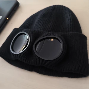 Svart mössa med glasögon - Cool svart stickad mössa med inbyggda glasögon framtill. Perfekt för att ge en unik touch till din outfit. Glasögonen är fastsatta och ger en futuristisk look.