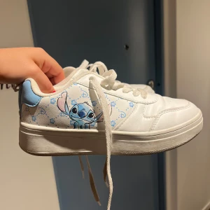 Vita sneakers med Stitch-motiv - Snygga vita sneakers med ett gulligt Stitch-motiv på sidan. Skorna har blå detaljer och klassisk snörning. Perfekta för dig som älskar Disney och vill ha en unik touch på dina sneakers.