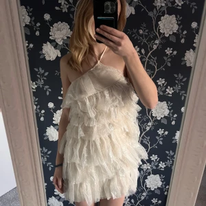 Glittrig halterneck klänning - Säljer en superfin glittrig klänning i ljus färg med volanger. Aldrig använd - lappen finns kvar. Den är axelbandslös och perfekt för festliga tillfällen. Klänningen är ganska kort, jag är 170 cm för referens.