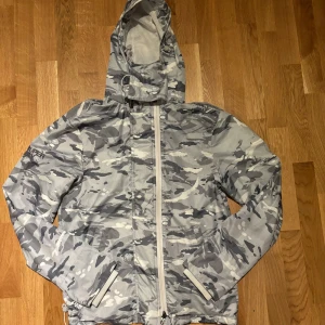 Windbreaker Superdry - Säljer en grå camouflagemönstrad vindjacka från Superdry. Jackan har en huva och dragkedja framtill. Perfekt för blåsiga dagar med sin stilrena design och praktiska fickor. Superdry-logga på ryggen och armen ger en extra touch. Storleken är S men passar mig på 185cm som brukar ha M-L. Den har 3 lager med dragkedjor så man kan välja själv hur tajt den ska sitta. Har även spännen längst ner man kan dra åt!