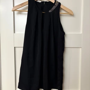 Svart ärmlös lång blus från Promod - Snygg svart ärmlös lång blus från Promod med dekorativa pärlor och stenar på axeln. Perfekt för en elegant look.