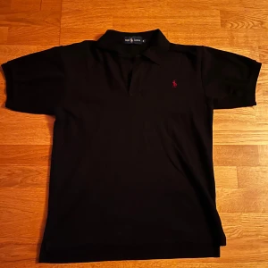 Svart pikétröja från Ralph Lauren - Säljer en klassisk svart pikétröja från Ralph Lauren med det ikoniska röda broderade logotypen på bröstet. Tröjan har korta ärmar och en stilren design som passar perfekt till många tillfällen. Storlek M