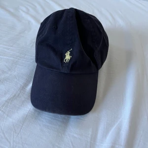 Ralph Lauren keps - Snygg mörkblå keps från Ralph Lauren med det klassiska broderade logotypen i gult framtill. Kepsen har justerbar rem baktill. Keps är i bra skick utan skador och defekter. Hör av er vid frågor. Mvh Eric