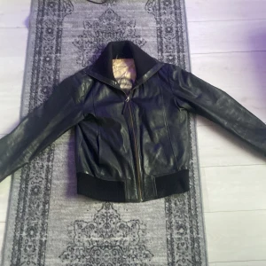 Svart skinnjacka  - Snygg svart skinnjacka med dragkedja och ribbade muddar. Jackan har en klassisk look med långärmade ärmar och en stilren design. Perfekt för en cool och tidlös stil. Från märket ”Rock and blue”