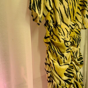 Zebramönstrad klänning med rynkdetaljer - Snygg zebramönstrad klänning i gult och svart med rynkdetaljer längs sidorna. Klänningen har korta ärmar och en figurnära passform som ger en trendig look. Perfekt för att sticka ut med ett djärvt mönster.