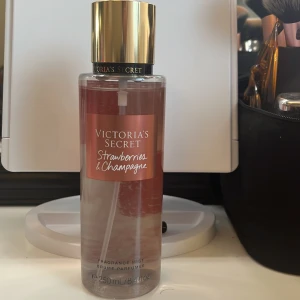 Victoria's Secret Strawberries & Champagne Fragrance Mist - En lyxig doftmist från Victoria's Secret med en fräsch och bubblig doft av vilda jordgubbar och mousserande champagne. Flaskan är elegant med en rosa etikett och guldfärgad kork. Perfekt för att ge en fruktig och festlig känsla.