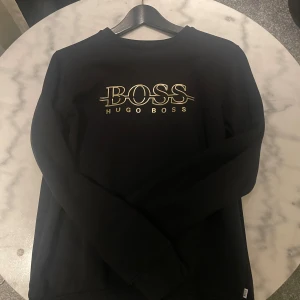 Svart tröja från Hugo Boss - Snygg svart tröja från Hugo Boss med guldigt tryck på bröstet. Tröjan har långa ärmar och en klassisk rund halsringning. Perfekt för en stilren look. finns defekt på trycket. 