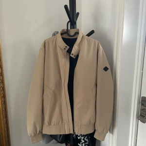 Beige Derek Poly Jacket från J.Lindeberg - Säljer en stilren beige Derek Poly Jacket från J.Lindeberg. Jackan har en krage med knappar och stängs med tvåvägsblixtlås. Den har två fickor fram. Vattenavstötande och PFC-fritt tyg med logotypemblem på vänster ärm. Nypris 3.000kr!