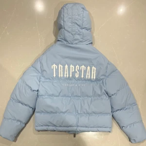 Ljusblå pufferjacka från Trapstar - Säljer en snygg ljusblå pufferjacka från Trapstar med broderad logga på ryggen. Jackan har en huva och dragkedja framtill. Perfekt för kyliga dagar!