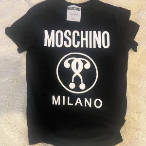 Svart t-shirt från Moschino - Snygg svart t-shirt från Moschino med stort vitt tryck på framsidan. Tillverkad i 100% bomull för en bekväm känsla. Perfekt för dig som vill ha en stilren och trendig look.