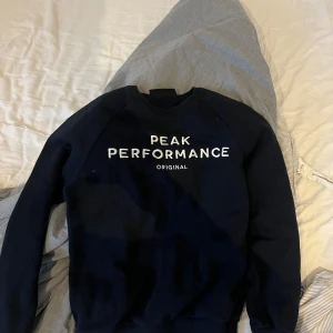 Svart sweatshirt från Peak Performance - Säljer en stilren svart sweatshirt från Peak Performance med vit text på bröstet. Tröjan har långa ärmar och en klassisk rund halsringning. Perfekt för en avslappnad look.