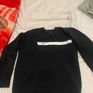 Svart tröja från Boss - Säljer en stilren svart tröja från Boss med en vit horisontell rand och texten 'BOSS' på bröstet. Tröjan har långa ärmar och en klassisk rund halsringning. Perfekt för en avslappnad och trendig look.
