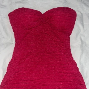 Röd/rosa axelbandslös topp - Snygg röd axelbandslös topp. Klänningen har en texturerad yta och en inre struktur för extra stöd. Perfekt för en kväll ute eller en speciell tillställning.