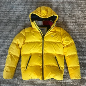 Gul pufferjacka från Tommy Jeans - Säljer en superfin gul pufferjacka från Tommy Jeans. Stlk M men passar ungdom/small. Den är perfekt för kalla dagar med sin varma vaddering och har en praktisk huva. Jackan har dragkedja och två fickor med dragkedjor. Den är i mycket bra skick och redo för vinterns äventyr! 💛