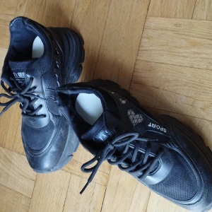 Svarta sneakers med tjock sula - Säljer ett par svarta sneakers i bra skick. De har en chunky sula och snörning framtill. Perfekta för en sportig look eller vardagsbruk. Märket 'Fashion Sport' syns tydligt på sidan och tungan av skorna. Passar bra till både jeans och träningskläder.