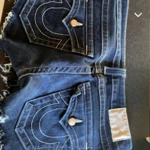 Mörkblå true religion shorts - Säljer ett par mörkblå jeansshorts med snygga fransar och coola fickdetaljer. Perfekta för sommardagar och festivaler! De har en klassisk femficksdesign och knappar i silver. Passar perfekt till en avslappnad stil.