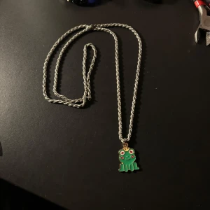 Sött halsband med groda - Det här är ett supergulligt halsband med en grön groda som hänge. Kedjan är tvinnad och ser ut att vara i silverfärg. Perfekt för att lägga till en lekfull touch till din outfit. Grodan har små rosa detaljer som gör den extra charmig. Passar perfekt för vardagsbruk eller som en rolig accessoar på fest!