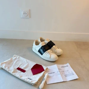 Valentino Garavani sneakers - Säljer ett par snygga Valentino Garavani sneakers i vitt med ett brett svart band över snörningen. Skorna är i mycket bra skick och kommer med originalpåse och extra skosnören. Perfekta för både vardag och fest! Skicket är 10/10 och de är aldrig använda. Fick dessa på min födelsedag förra året och har inte gillat dom sen dess. Köpta på NK!!!