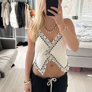 Vit broderad topp med snörning - Säljer en superfin vit topp med svart broderi och snörning i ryggen. Toppen är ärmlös och har en unik omlott-design framtill. Helt oanvänd med lappen kvar!