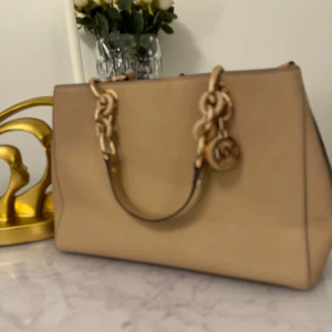 MK Beige handväska med kedjedetaljer - MK snygg beige handväska i mycket bra skick med guldfärgade kedjedetaljer och logotyp. Väskan har flera fack och dragkedja inuti, perfekt för att hålla ordning på dina saker. Stilren och passar till både vardag och fest.