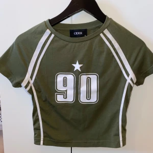 Olivgrön croppad t-shirt från Cider - Säljer en olivgrön croppad t-shirt från Cider med vita detaljer och siffran 90 tryckt på framsidan. T-shirten har korta ärmar och en stjärna ovanför siffrorna. Perfekt för en avslappnad stil!