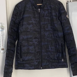 Camouflage mönstrad jacka från Moncler. Storlek L - Säljer en snygg och stilren pufferjacka från Moncler i blått och svart camouflage. Jackan har en dragkedja framtill och långa ärmar. Perfekt för höst och vår när du vill hålla stilen. Den är i mycket bra skick och redo för nya äventyr!