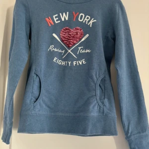 Blå hoodie från Tommy Hilfiger - Säljer en snygg blå hoodie från Tommy Hilfiger med texten 'New York Rowing Team Eighty Five' och ett hjärta i röda paljetter på framsidan. Den har en mysig huva och praktiska fickor. Perfekt för en avslappnad stil! 💙Köparen betalar frakt. 