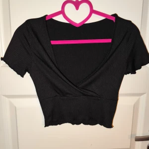 Svart ribbad omlottopp - Säljer en snygg svart ribbad omlottopp med korta ärmar. Toppen har en croppad passform och är perfekt för både vardag och fest. Den är i mycket bra skick och passar till många olika outfits.