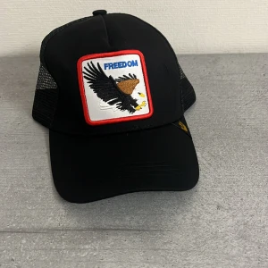 Svart keps med örn och texten 'Freedom' - Snygg svart keps med ett broderat märke av en örn och texten 'Freedom' på framsidan. Kepsen har ett nätmaterial på sidorna och justerbar passform baktill. Perfekt för en avslappnad stil eller sommaräventyr. Ny och oanvänd 